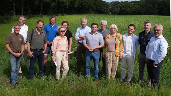 Groepsfoto van dertien mensen die buiten op een grasveld staan met een groene akker en bomen op de achtergrond, onder een blauwe lucht met wolken.