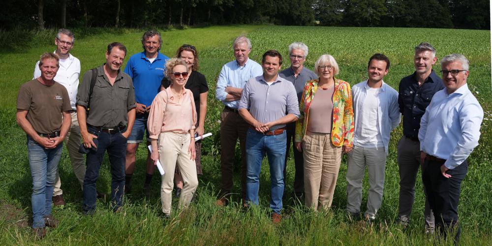 Groepsfoto van dertien mensen die buiten op een grasveld staan met een groene akker en bomen op de achtergrond, onder een blauwe lucht met wolken.